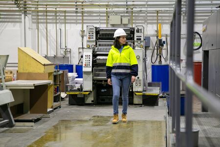 Confident female printing worker walking at factory. Mature woman in yellow hard hat walking near...