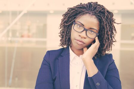 Pensive female office worker wearing glasses, posing outside. Young African American business woman standing at glass wall, touching ear, looking at camera. Businesswoman conceptの写真素材