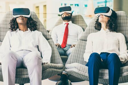 Group of coworkers in VR headsets. Front view of multiethnic businesswomen and businessman sitting in chairs and using virtual reality headsets. Technology conceptの写真素材