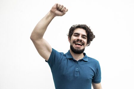 Happy excited guy making winner gesture, enjoying success. Handsome bearded young man in blue casual t-shirt posing isolated over white background. Joy or euphoria conceptの写真素材