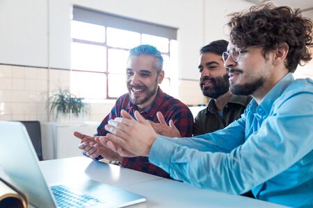 Group of coworkers watching online training or webinar on laptop, applauding speaker. Business colleagues in casual meeting in contemporary office space. Online training conceptの写真素材