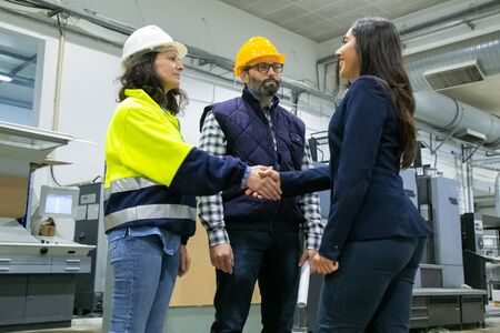 Positive businesswoman and engineers meeting onsite and shaking hands. Man and woman in hardhat, uniform and business suit standing on plant floor. Business and industry conceptの写真素材