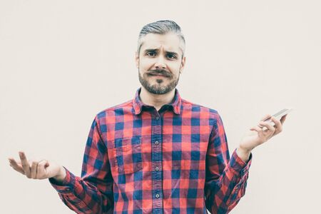 Doubtful man holding smartphone and looking at camera. Front view of uncertain bearded man in...