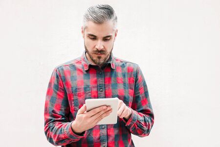 Serious man using tablet pc. Front view of focused bearded man in checkered shirt using tablet computer on grey background. Wireless technology conceptの写真素材