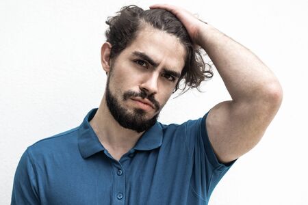 Pensive attractive guy touching head, doing up hair, looking at camera. Handsome bearded young man...