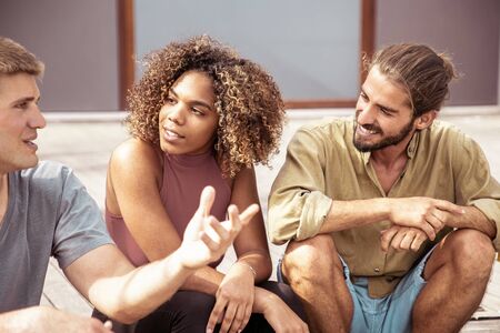 Happy young friends talking outdoors. Cropped shot of cheerful young multiethnic friends sitting on wooden surface and talking on street. Communication conceptの写真素材