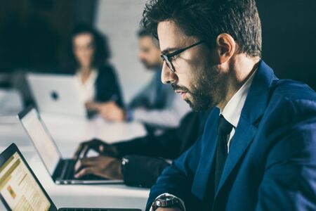 Serious businessman sitting at table and looking at laptop. Side view of focused man in eyeglasses...