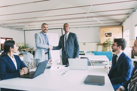 Two businessmen shaking hands in office. Smiling colleagues standing and looking at camera. Business...