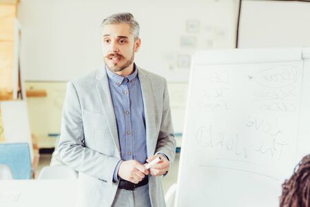 Business coach talking while standing near whiteboard. Confident Caucasian manager doing...