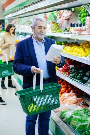 Mature man buying vegetables in supermarket. Bearded man in eyeglasses holding basket and choosing fresh vegetables in grocery store. Shopping conceptの写真素材