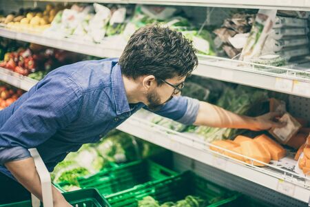 Serious young man shopping in grocery store. Side view of focused young man in eyeglasses choosing fresh vegetables in supermarket. Shopping conceptの写真素材