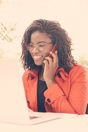 Smiling young woman talking on phone and looking at laptop. Cheerful African American lady in...
