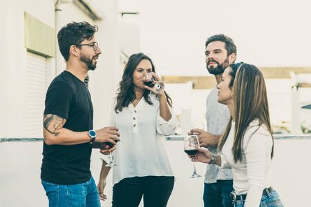 Smiling young people drinking red wine and talking. Cheerful friends holding glasses with wine at balcony. Leisure, friendship conceptの写真素材