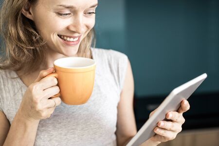 Closeup portrait of smiling young beautiful woman drinking tea from cup and using tablet computer. Morning news concept.の写真素材