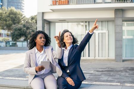 Businesswomen using tablet pc and pointing up. Multiethnic businesswomen sitting together outdoor, using digital tablet and pointing away with fingers. Wireless technology conceptの写真素材