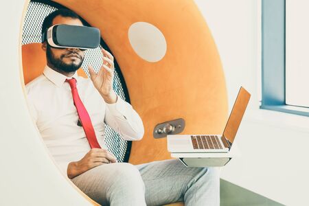 Businessman using VR headset and laptop. Serious young African American businessman sitting in spherical chair and using virtual reality headset in office. Technology conceptの写真素材