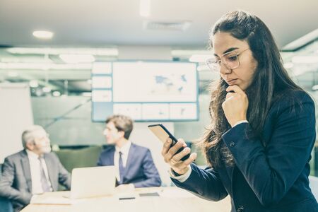 Thoughtful businesswoman sitting on table and looking at phone. Focused young woman in eyeglasses using smartphone. Business, technology conceptの写真素材