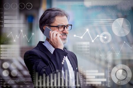 Businessman calling on smartphone and virtual statistic graphics. Closeup portrait of smiling middle-aged handsome man looking away and talking on phone outdoorsの写真素材