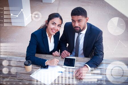 People with tablet, document and virtual statistic graphics. Man and woman wearing formal clothes, looking at camera and sitting at office desk. Business work concept. High angle view front view.の写真素材