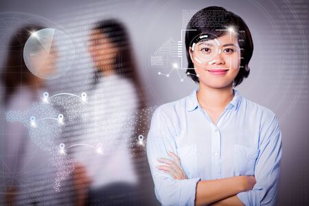Young Asian businesswoman and virtual identification graphics. Portrait of happy woman standing with folded arms, looking at camera and smiling, her colleagues talking in backgroundの写真素材