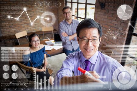 Asian expert giving presentation and virtual statistic graphics. Closeup of smiling senior businessman writing on whiteboard in loft conference room in background. High angle view.の写真素材