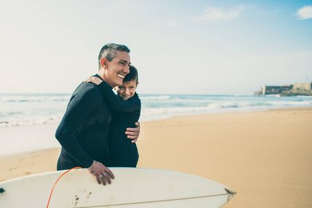 Father and son in wetsuits hugging on beach. Cheerful father and son hugging while spending time together with surfboard on beach. Water sport conceptの写真素材