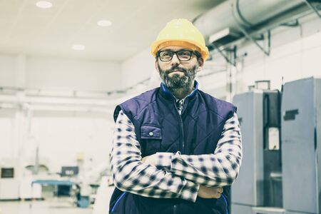 Indoor portrait of positive confident factory worker. Bearded middle aged man in glasses, hardhat...
