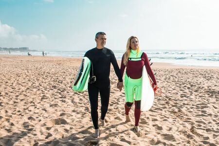 Couple of surfers walking on sandy beach. Front view of middle aged man and woman in wetsuits...