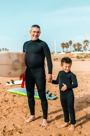 Father and son in wetsuits smiling at camera. Cheerful middle aged father holding surfboard and standing with cute little son on sea coast. Water sport conceptの写真素材
