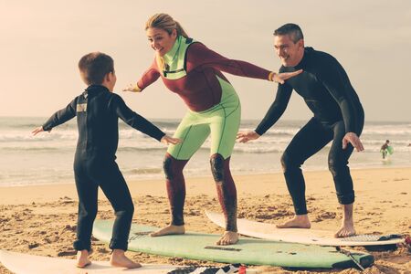 Family sanding on surfboards on beach. Parents and little son in wetsuits standing on boards and learning surfing on sandy sea coast. Water sport conceptの写真素材