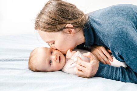 Mother kissing baby daughter in bedroom. Side view of happy young mother hugging and kissing adorable infant child at home. Parenthood conceptの写真素材