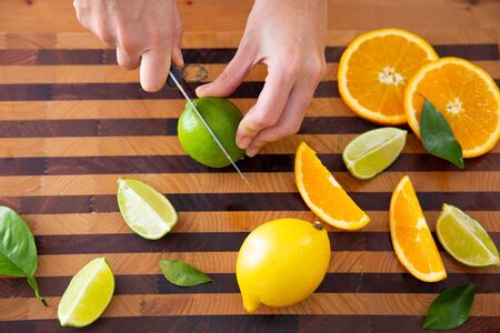 Woman holding and cutting lime in two pieces with knife. Sliced orange, lemon and leaves on wooden board. Top view. Studio shot. Nutrition and vegetarian conceptの写真素材