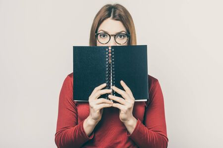 Female college student hiding behind notebook. Young woman in eyeglasses holding textbook near face and looking at camera isolated on white background. Education conceptの写真素材