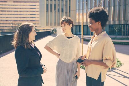 Three women with smartphones standing on street. Cheerful female friends looking at phone and talking. Technology, leisure conceptの写真素材