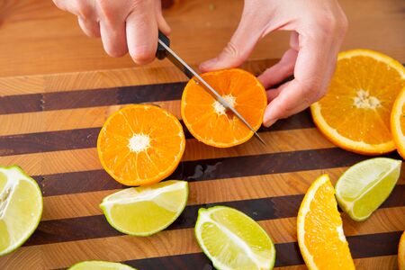 Female hands holding and cutting mandarin in four pieces with knife. Sliced orange and lime on wooden board. Side view. Studio shot. Nutrition and vegetarian conceptの写真素材