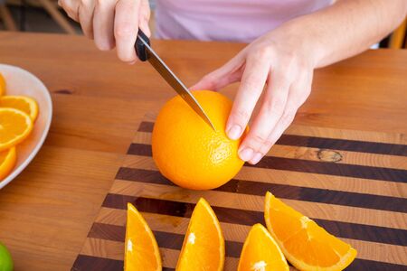 Female hands holding knife and cutting orange on wooden board. White plate with sliced citrus...