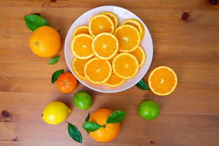 Big white ceramic plate with sliced oranges. Fresh mandarins, limes, lemon and citrus with leaves on wooden desk near bowl. Top view. Studio shot. Nutrition and vegetarian conceptの写真素材