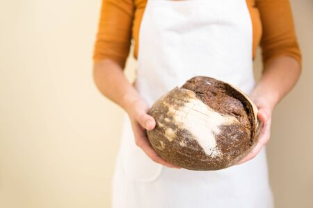 Baker wearing apron, holding loaf of black rye bread. Closeup of person holding food. Baking or...