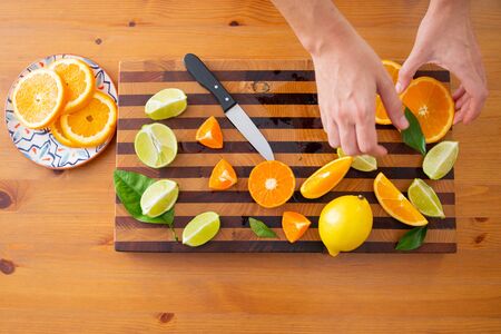 Hands taking citrus fruits from table. Sliced orange in ceramic plate. Lime, lemon, citrus leaves and knife on wooden board. Top view. Studio shot. Nutrition and vegetarian conceptの写真素材