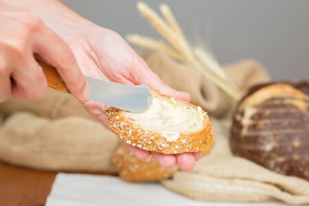 Rustic breakfast cooking. Hands of woman spreading butter onto slice of baguette with knife. Closeup...