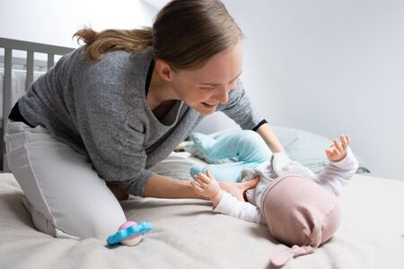 Cheerful happy mom caressing baby daughter in bed. Mother and little child staying at home. Child...