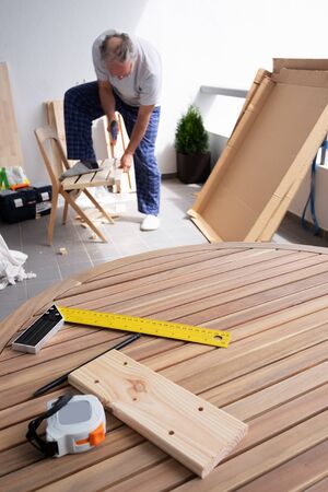 Caucasian senior man sawing wood during pandemic isolation. Construction rulers laying on round table. House improving and home decoration during quarantine conceptの写真素材