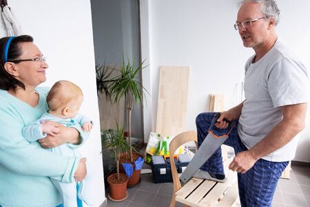 Smiling woman with baby talking with grey-haired craftsman. Granddad working with wood at balcony. House improving, DIY and home decoration during quarantine conceptの写真素材