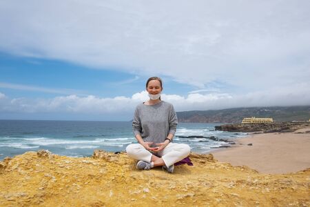 Positive young woman with closed eyes in face mask, practicing yoga and meditation on ocean beach. Front view. Staying well during outbreak conceptの写真素材