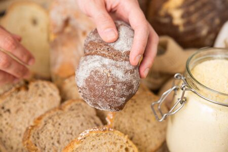Hands of woman holding rye bun over heap of toasts and freshly baked loafs. Closeup shot, side view....