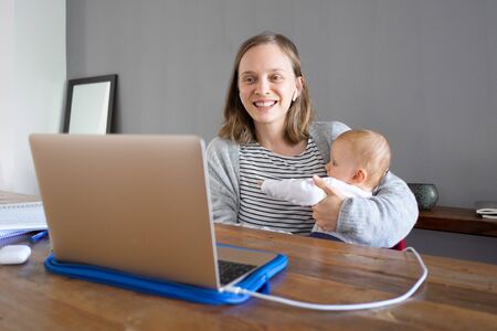 Smiling mother with daughter calling to friend via laptop. Young woman holding cute infant and chatting via computer. Online connection and video call worldwide at home conceptの写真素材