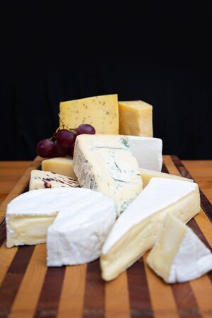 Heap of soft, blue and spicy cheeses on wooden chopping board against dark wall background. Closeup,...