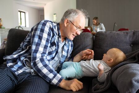 Grey-haired grandpa in glasses playing with baby girl on sofa. Concentrated mom out-of-focus working...