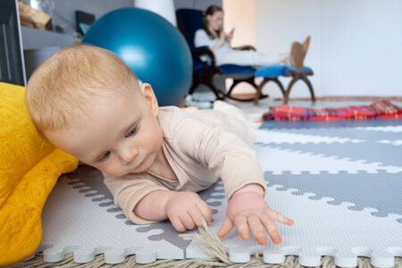 Sweet red haired baby girl trying to crawl on floor. Mom using smartphone in background. Childhood...