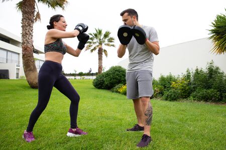 Positive woman practicing fighting skills with trainer. Man and woman boxing on grass. Outdoor fitness workout and physical activity concept.の写真素材